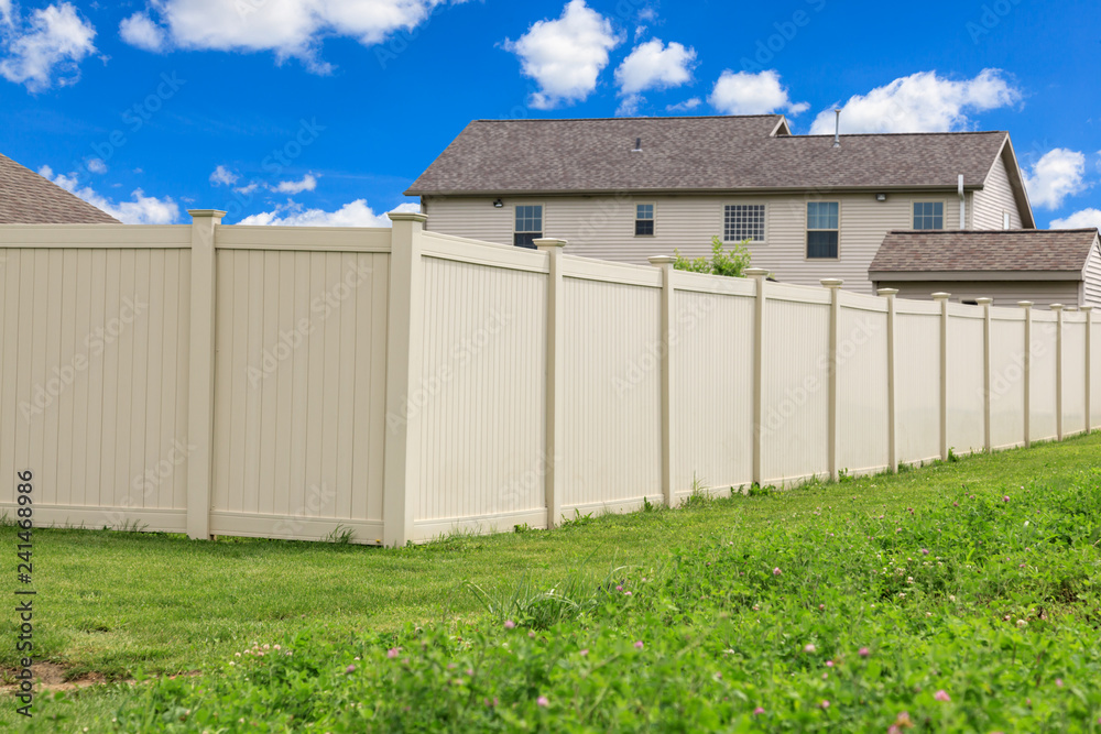 Tan colored vinyl fence surrounding a homes backyard. Grass and blue sky in the background.