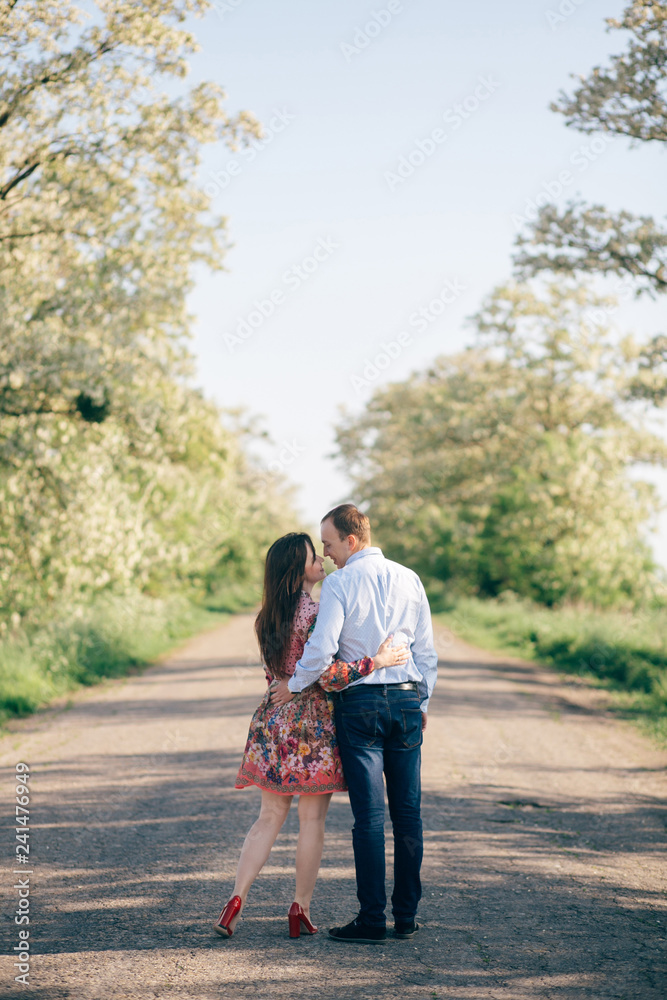 Beautiful young couple holding hands and walking on road in sunshine among spring field and trees. Happy family in love relaxing  in sunlight. Romantic joyful moments. Space for text