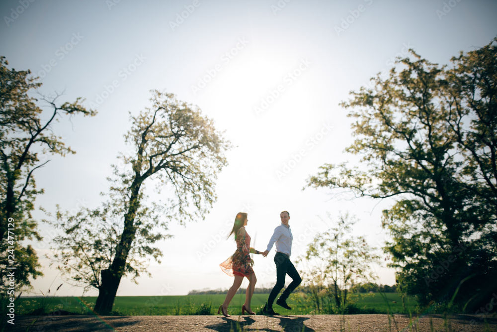 Beautiful young couple holding hands and running in sunshine in spring field and trees. Silhouettes of happy family having fun  in sunlight. Romantic joyful moments. Space for text