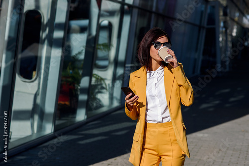 Caucasian young business woman using smart phone and drinks coffee outside near the office centre. Attractive woman scrolling pages, tapping on touchscreen, checking news and financial charts.