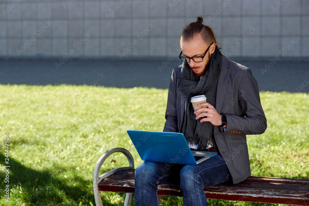 Handsome caucasian businessman with a beard sitting at the wooden bench with laptop and cup of coffee. Young male student sitting on the bench in the park and typing the text on the laptop.