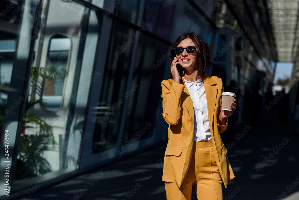 Fototapeta premium Smiling business woman in yellow suit walking near business centre at the city, communicates via smartphone and drinks coffee. Business concept, communication, banker, manager and office worker.