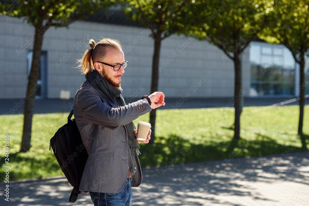 Fototapeta premium Young successful businessman in a grey jacket with scarf walks near the business centre, drinks coffee and looking on the watches. Concept of business, banker, manager.