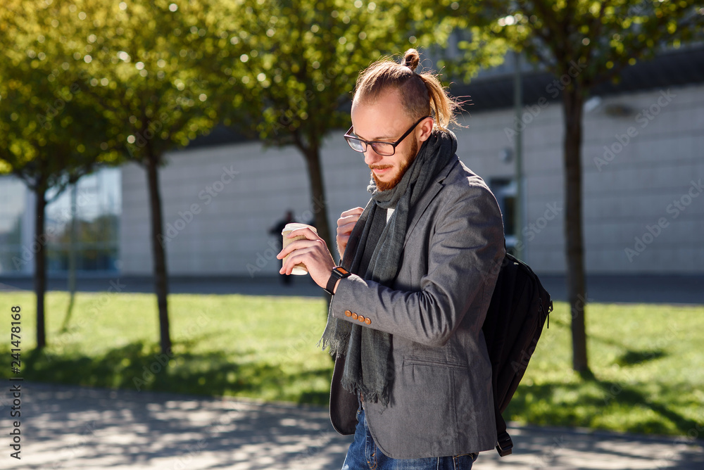 Young successful businessman in a grey jacket with scarf walks near the business centre, drinks coffee and looking on the watches. Concept of business, banker, manager. Young successful businessman in a grey jacket with scarf walks near the business centre, drinks coffee and looking on the watches. Concept of business, banker, manager.