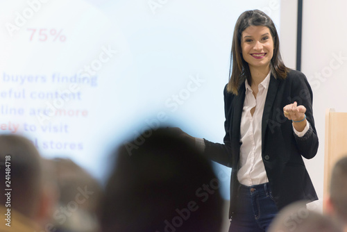Pretty young businesswoman, teacher or mentor coach speaking to young students in audience at training seminar, female business leader speaker talking at meeting.