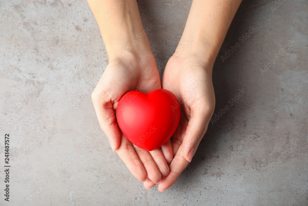 Fototapeta premium Woman holding red heart on gray table, top view. Cardiology concept