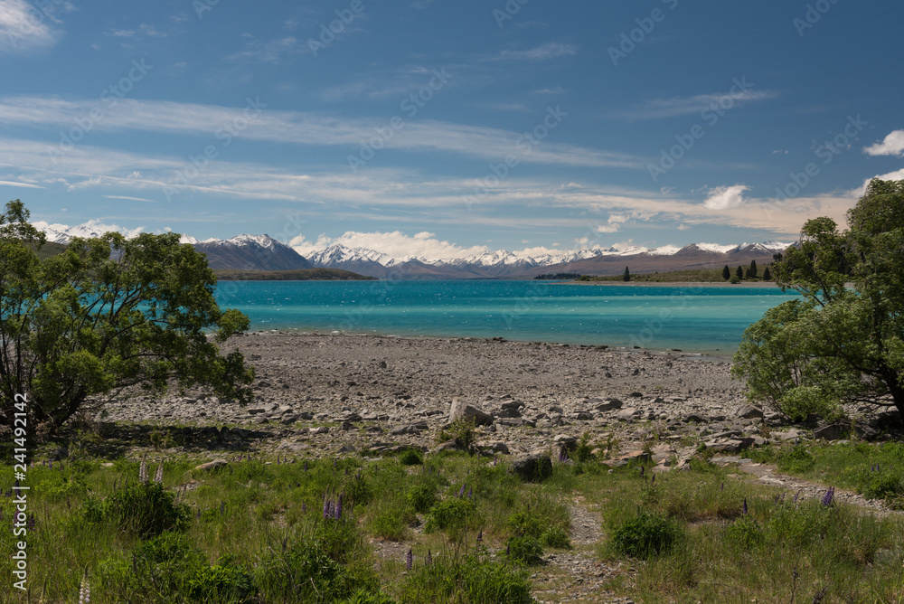 The view across a low Lake Tekapo with a stoney beach in the foreground and the snow capped Southern Alps in the background. In the Mackenzie Basin, New Zealand.