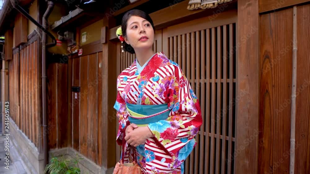 elegant local woman in flower pattern kimono standing in front of wooden home waiting for friends join festival together. young japanese lady in traditional costume looking around carrying purse. elegant local woman in flower pattern kimono standing in front of wooden home waiting for friends join festival together. young japanese lady in traditional costume looking around carrying purse.