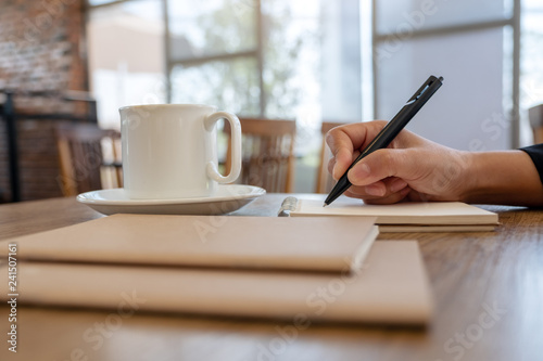Closeup image of a hand writing on blank notebook with coffee cup on table in cafe