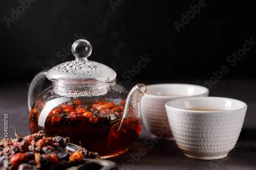Glass teapot with vitamin drink from hips and two bowls. Rosehip tea. Vitamin drink on a dark background.