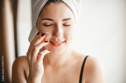 Young beautiful woman is smiling and putting cream on her face. Cozy homely atmosphere