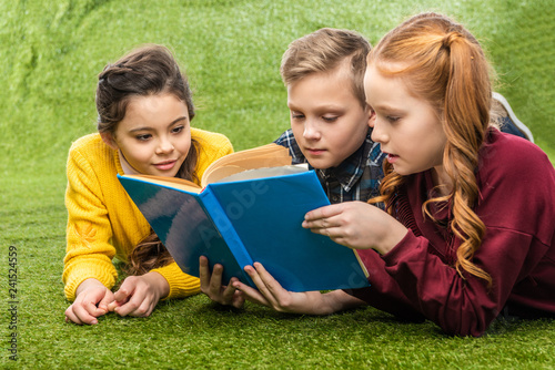 cute preteen schoolkids lying on green lawn and reading book