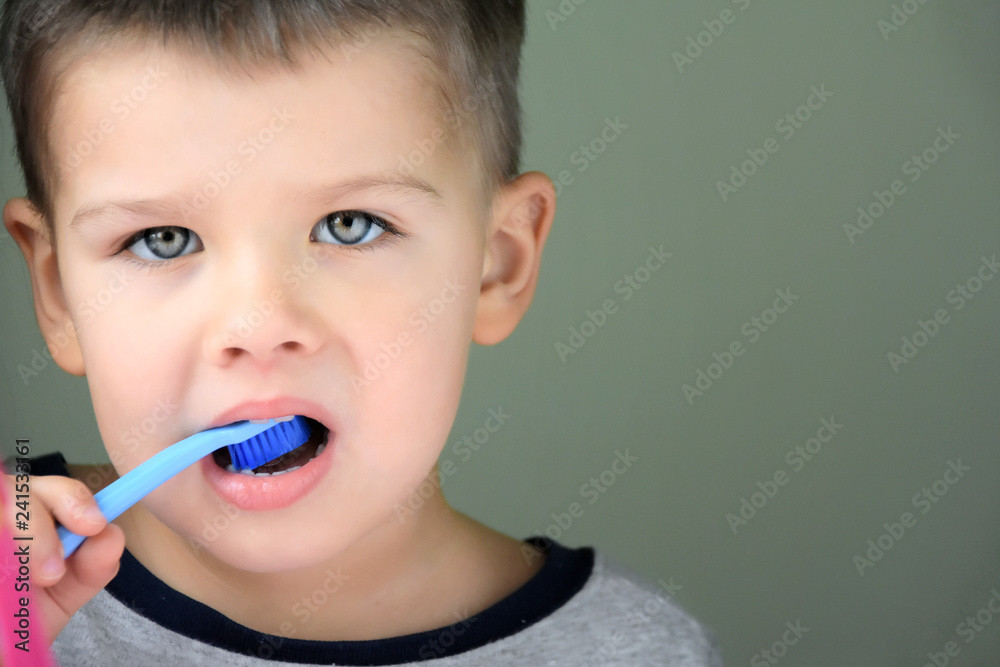 Cute smiling caucasian white boy with blue eyes brushing his teeth with colorful toothbrush on green background with blurred toothbrushes on front. Beautiful toddler boy doing morning hygiene. Cute smiling caucasian white boy with blue eyes brushing his teeth with colorful toothbrush on green background with blurred toothbrushes on front. Beautiful toddler boy doing morning hygiene.