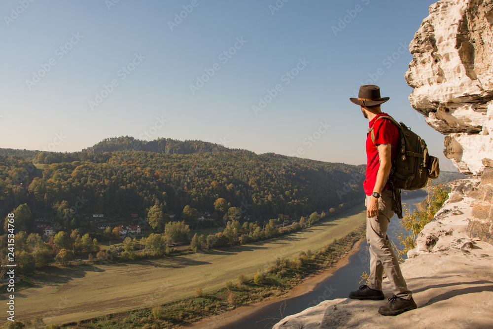 Young bearded male traveler in hat stand on the cliff in sunny day 