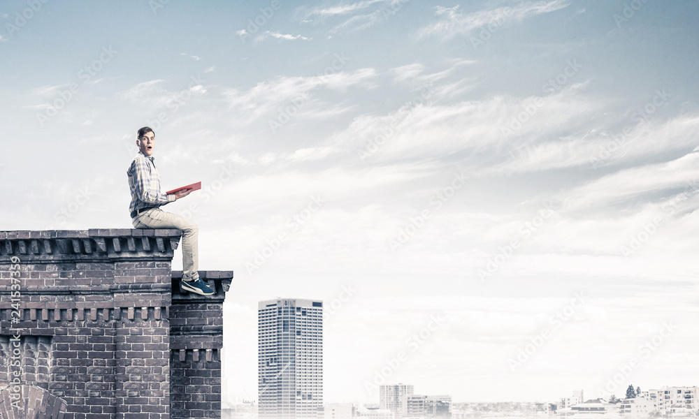 Man on roof edge reading book and cityscape at background Man on roof edge reading book and cityscape at background