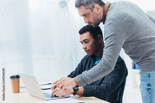 bearded mentor helping young colleague working with laptop in office photo