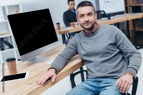 handsome man looking at camera while sitting at table with desktop computer and digital tablet in office
