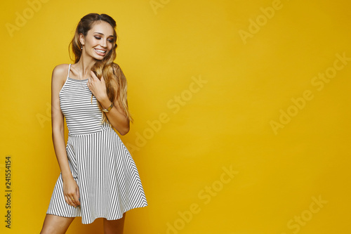 Fashionable and sexy blonde model girl with the attractive smile and with gentle makeup, in white striped dress adjusting her hairstyle and posing at the yellow background, isolated