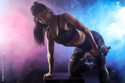 Sporty muscular woman with dumbbell in smoke on dark background