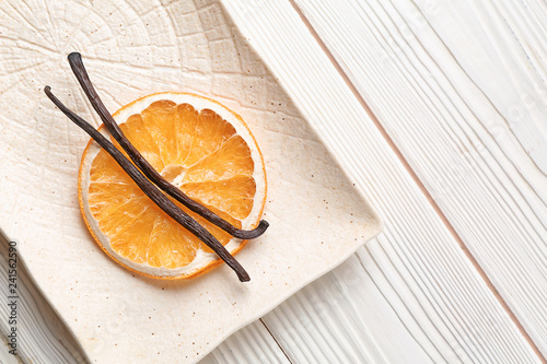 Plate with dry orange slice and aromatic vanilla sticks on white wooden background