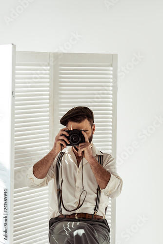 bearded man sitting near white room divider taking pictures with vintage film camera
