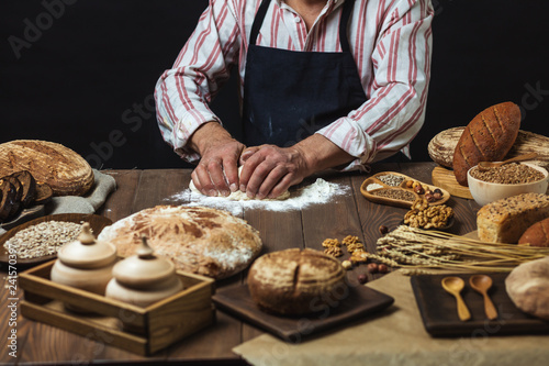 Chef or baker, dressed in black apron, preparing a portion of fresh dough in rural bakery, kneading the pastry surrounded by rustic organic loaf of bread.