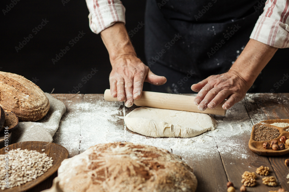 Chef or baker, dressed in black apron, preparing a portion of fresh dough in rural bakery, kneading the pastry surrounded by rustic organic loaf of bread.