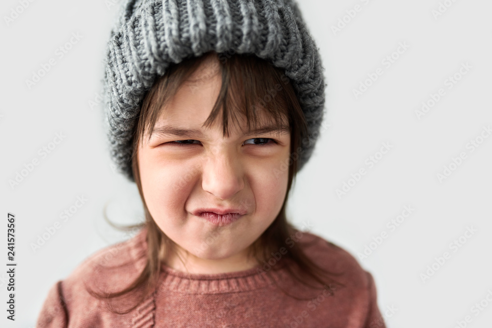 Studio closeup portrait of cute unhappy little girl with grumpy emotion in the winter warm gray hat, wearing sweater isolated on a white studio background. Studio closeup portrait of cute unhappy little girl with grumpy emotion in the winter warm gray hat, wearing sweater isolated on a white studio background.