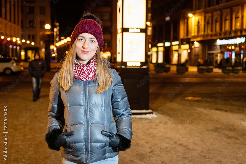 Winter portrait of happy young woman walking in snowy city decorated for Christmas and New Year holidays. Christmas lights on background. - Image