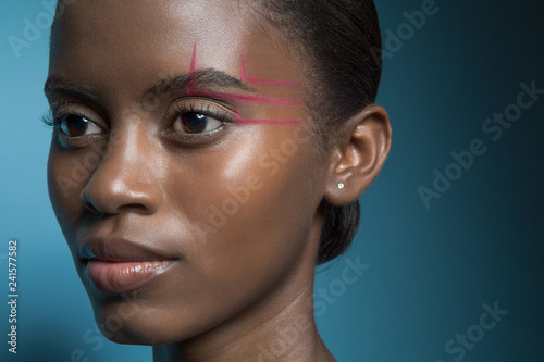 Portrait of a beautiful, young, black woman wearing graphic make up in front of a blue background.