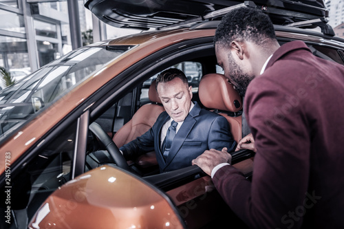 Bearded sales manager of car salon standing near client sitting in car