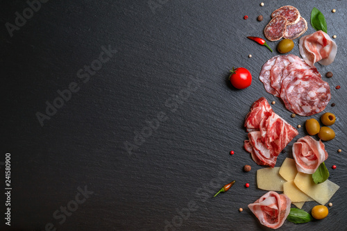 flat lay of antipasti with salami, cheeses, prosciutto, ham, olives, greens, tomato on black slate background, copy space