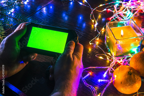 Man hand holding blank green screen mobile smart phone with Christmas light decorated background, home use, wooden table top view, play game, watching video
