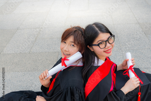 Portrait image of Two teenage girl Smiling bright, wearing a graduation gwon On graduation day, sit back against each other on the floor, to education concept. photo