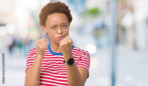 Beautiful young african american woman wearing glasses over isolated background Ready to fight with fist defense gesture, angry and upset face, afraid of problem © Krakenimages.com