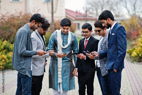 Group of six south asian indian mans in traditional, casual and business wear standing and looking at mobile phones together.