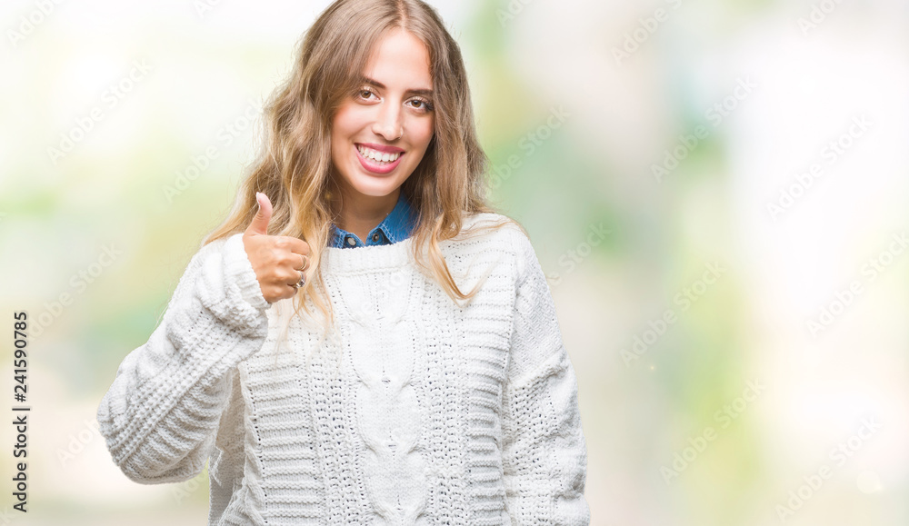 Beautiful young blonde woman wearing winter sweater over isolated background doing happy thumbs up gesture with hand. Approving expression looking at the camera with showing success.