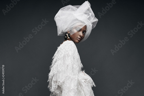 Fashion portrait of woman in white lace dress with white veil head tie and big earring. Studio, grey background photo