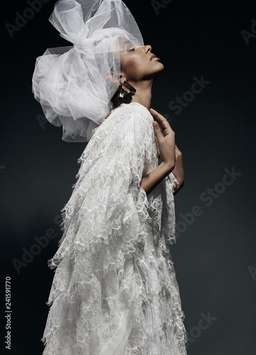 Fashion portrait of woman in white lace dress with white veil head tie and big earring. Studio, grey background photo