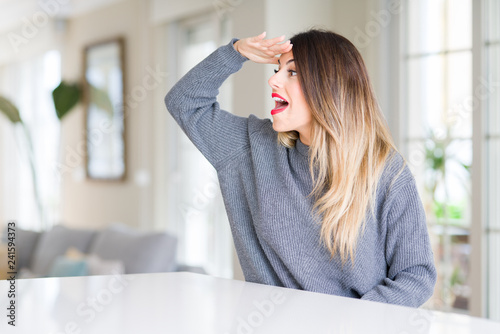 Young beautiful woman wearing winter sweater at home very happy and smiling looking far away with hand over head. Searching concept.
