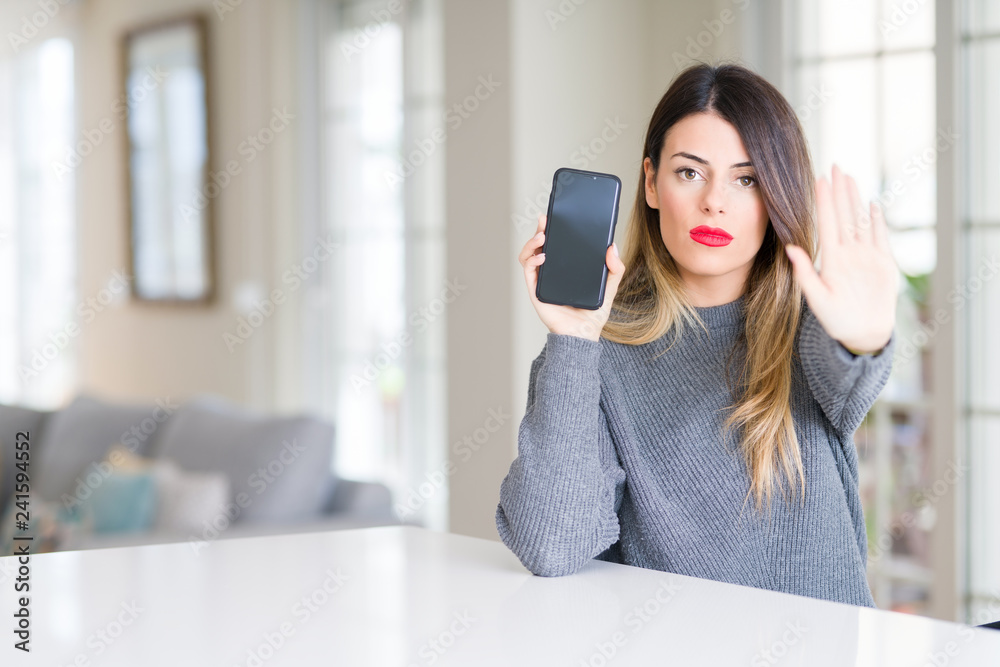 Young beautiful woman showing smartphone screen at home with open hand doing stop sign with serious and confident expression, defense gesture Young beautiful woman showing smartphone screen at home with open hand doing stop sign with serious and confident expression, defense gesture