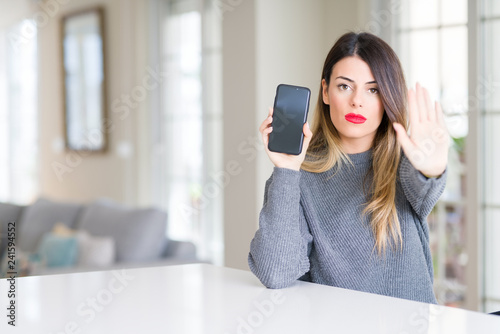 Young beautiful woman showing smartphone screen at home with open hand doing stop sign with serious and confident expression, defense gesture