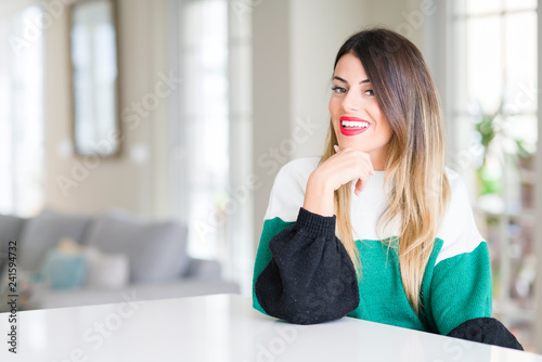 Young beautiful woman wearing winter sweater at home looking confident at the camera with smile with crossed arms and hand raised on chin. Thinking positive.