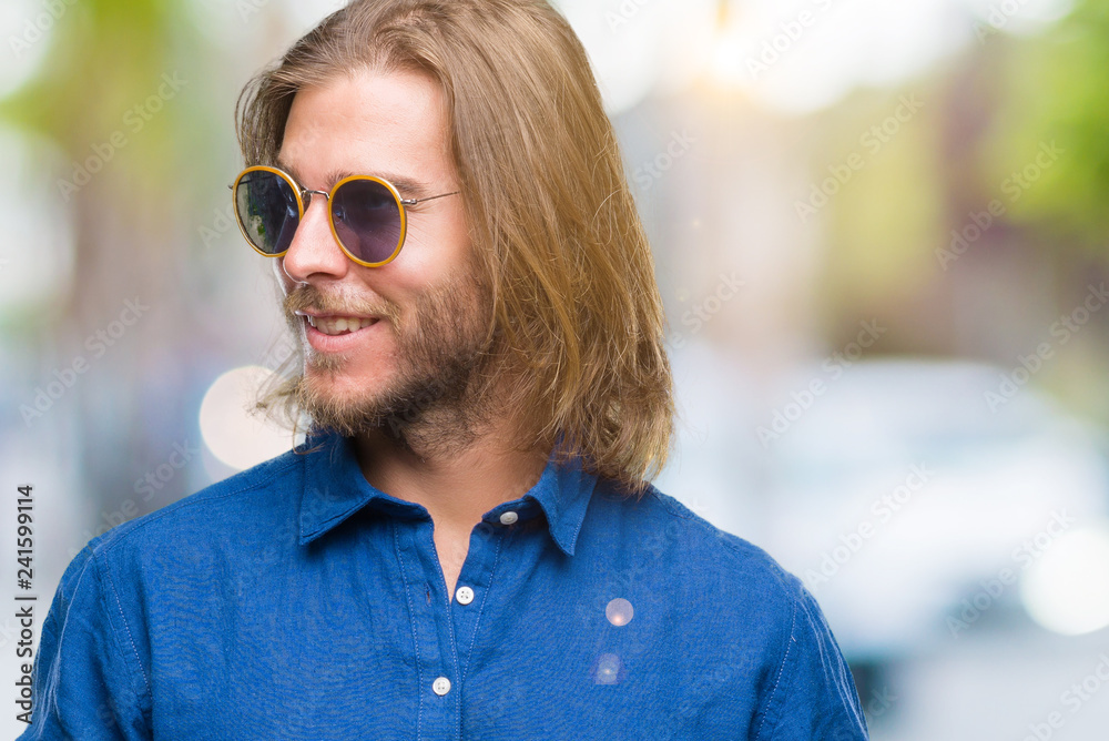 Young handsome man with long hair wearing sunglasses over isolated background looking away to side with smile on face, natural expression. Laughing confident.