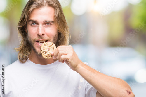 Young handsome man with long hair eating chocolate cooky over isolated background with a confident expression on smart face thinking serious