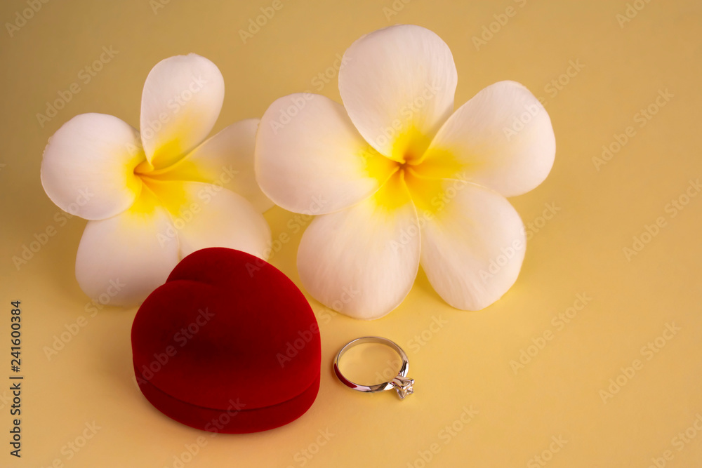 Elegant diamond engagement ring and red heart shaped box with tropical white flowers (plumeria) on yellow background. Concept for greeting card, postcard. Copy space. Elegant diamond engagement ring and red heart shaped box with tropical white flowers (plumeria) on yellow background. Concept for greeting card, postcard. Copy space.