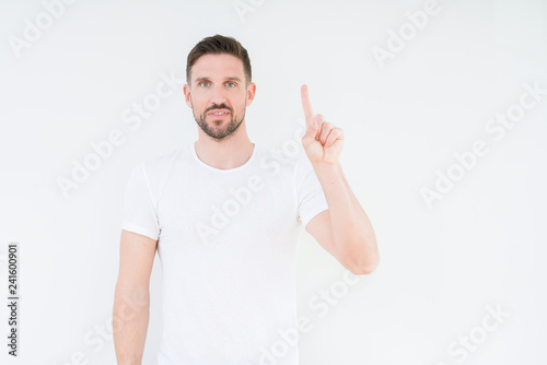 Young handsome man wearing casual white t-shirt over isolated background showing and pointing up with finger number one while smiling confident and happy.