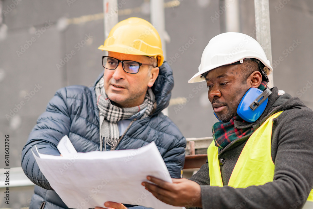 Civil engineers with hardhat and yellow jacket checking technical drawings and office blueprints among scaffolding on construction site Civil engineers with hardhat and yellow jacket checking technical drawings and office blueprints among scaffolding on construction site