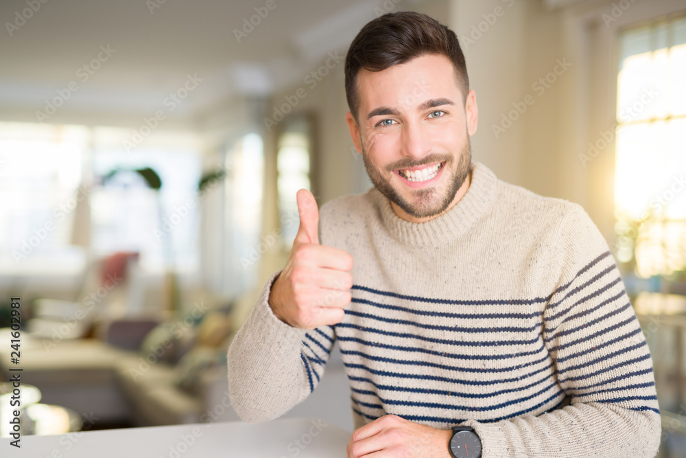 Young handsome man at home doing happy thumbs up gesture with hand. Approving expression looking at the camera with showing success. Young handsome man at home doing happy thumbs up gesture with hand. Approving expression looking at the camera with showing success.