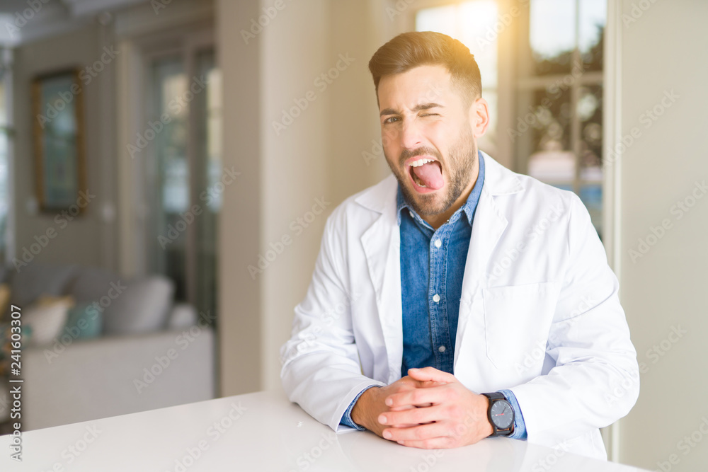 Young handsome doctor man at the clinic sticking tongue out happy with funny expression. Emotion concept.